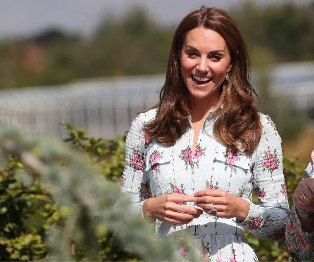 Smiling woman in a floral dress at a garden, likely at a festival, surrounded by greenery.