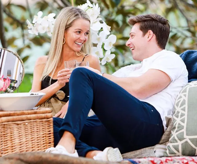 A couple sitting outdoors, smiling and clinking glasses, surrounded by cushions and flowers.