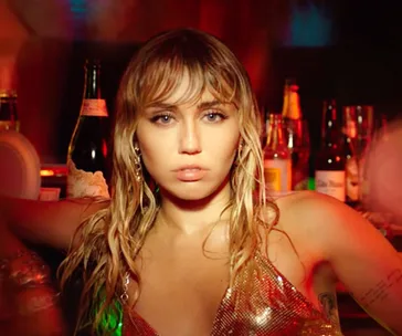 A woman with wet hair in a shiny gold top stands confidently in front of a bar with various bottles.