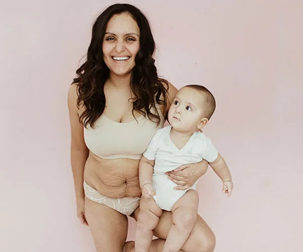 Smiling woman in underwear holding a baby, promoting body positivity against a pastel background.