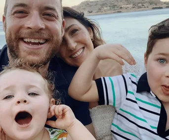 A family of four smiling by the seaside, with adults and two young children posing playfully for the camera.