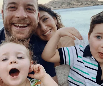 A family of four smiling by the seaside, with adults and two young children posing playfully for the camera.