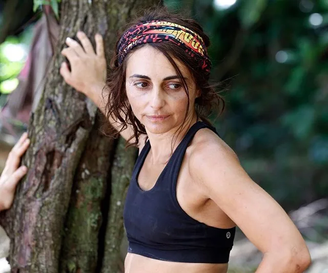 Person wearing a headband and black tank top stands by a tree in a jungle setting.