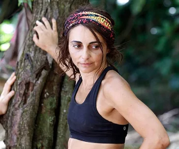 Person wearing a headband and black tank top stands by a tree in a jungle setting.