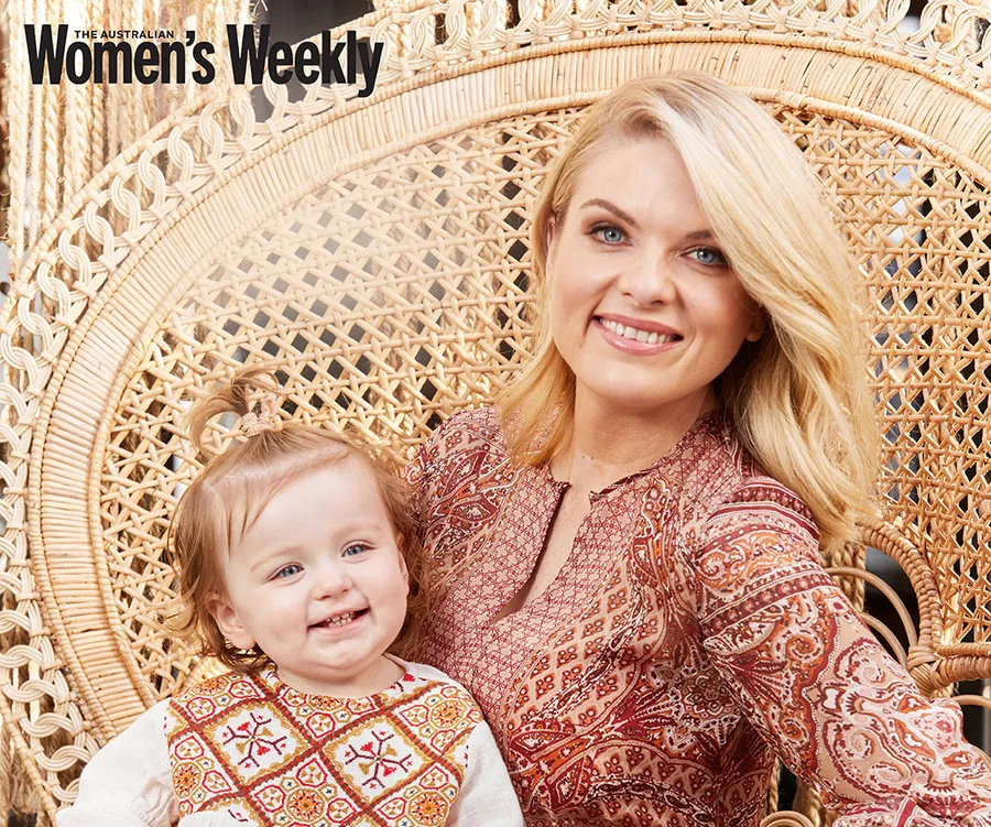 A woman and young child sit on a wicker chair, smiling. The woman has blonde hair and the child is wearing an embroidered outfit.