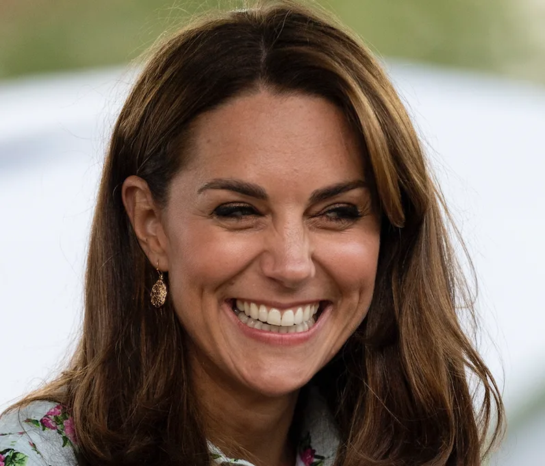 A woman with long brown hair smiling broadly, wearing earrings, in an outdoor setting.