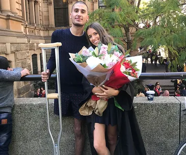 Young couple smiling, the man with crutches, the woman holding a bouquet, standing outside a historic building.