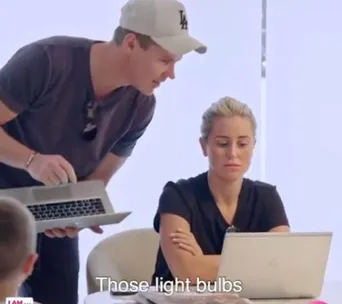 Man in cap talks while showing a laptop to a seated woman who looks at her own laptop; subtitle reads "Those light bulbs."