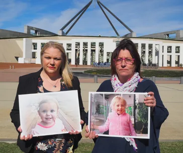 Women holding photos of a girl outside a large government building under a clear blue sky.
