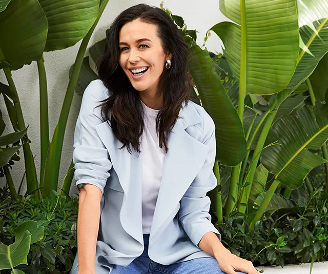 A woman in a light blue blazer sitting and smiling in a garden setting with large green leaves in the background.