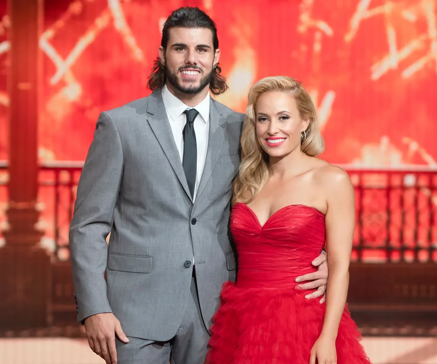 Couple dressed elegantly; man in a gray suit, woman in a red dress, standing with a warm backdrop.