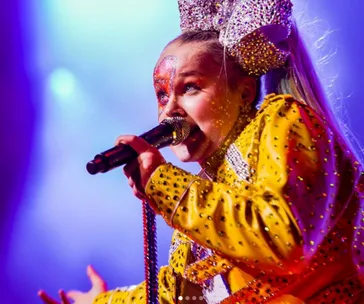JoJo Siwa performs energetically in a sparkly yellow outfit with a large bow, holding a microphone, on stage.