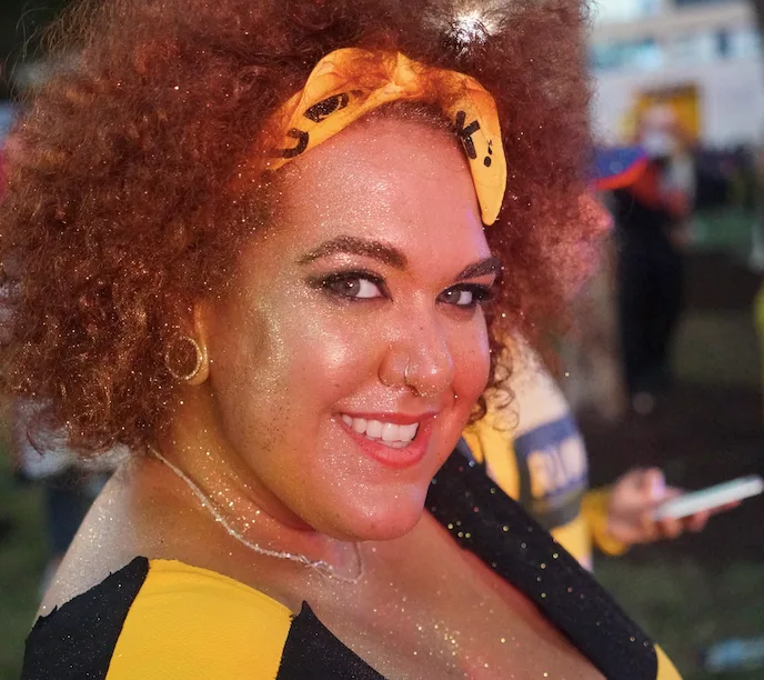A person with curly hair, sparkling makeup, and a yellow headband smiles at a festive outdoor event.