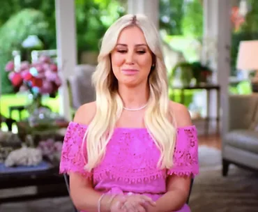 Blonde woman in a bright pink dress sits indoors, smiling softly, with a blurred garden visible through large windows.