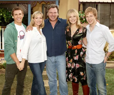Cast members of "Packed to the Rafters" pose together outdoors, smiling at the camera.