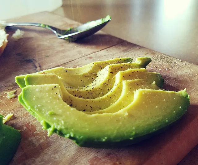 Sliced avocado with black pepper on a wooden cutting board next to a spoon.
