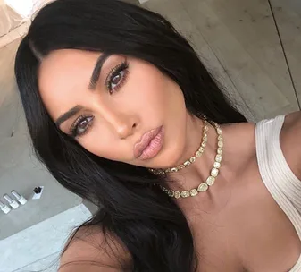 A person with long dark hair and contour makeup taking a selfie, wearing a layered necklace and white top.