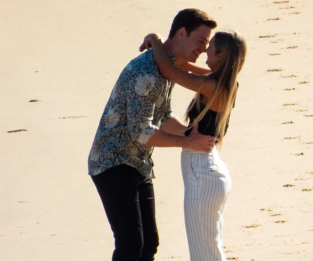 A couple embracing on a sunny beach, footprints in the sand behind them.