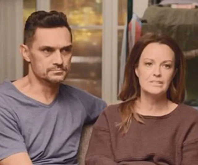 A couple sits indoors, looking serious and concerned, with a window and clothes in the background.