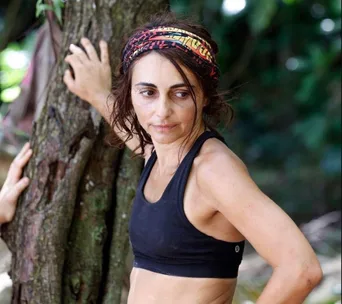 Woman in athletic wear and headband, leaning against a tree in a forest setting, looking contemplative.
