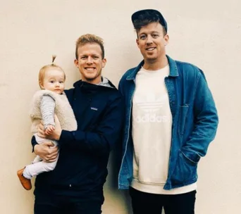 "Two men smiling, one holding a baby, standing in front of a plain wall."