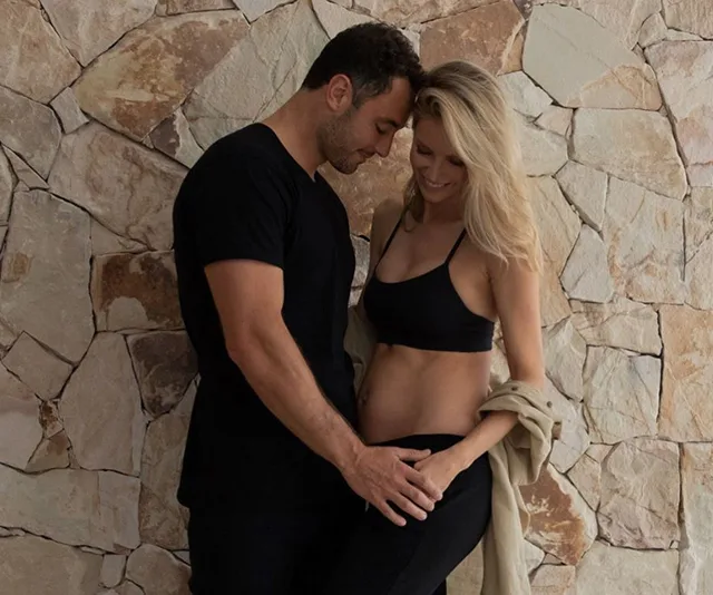 A couple embraces affectionately against a stone wall, with hands gently on a pregnant belly.