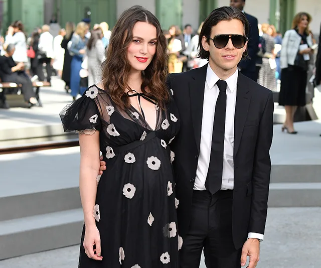 A woman in a black floral dress and a man in a suit with sunglasses pose together outdoors.