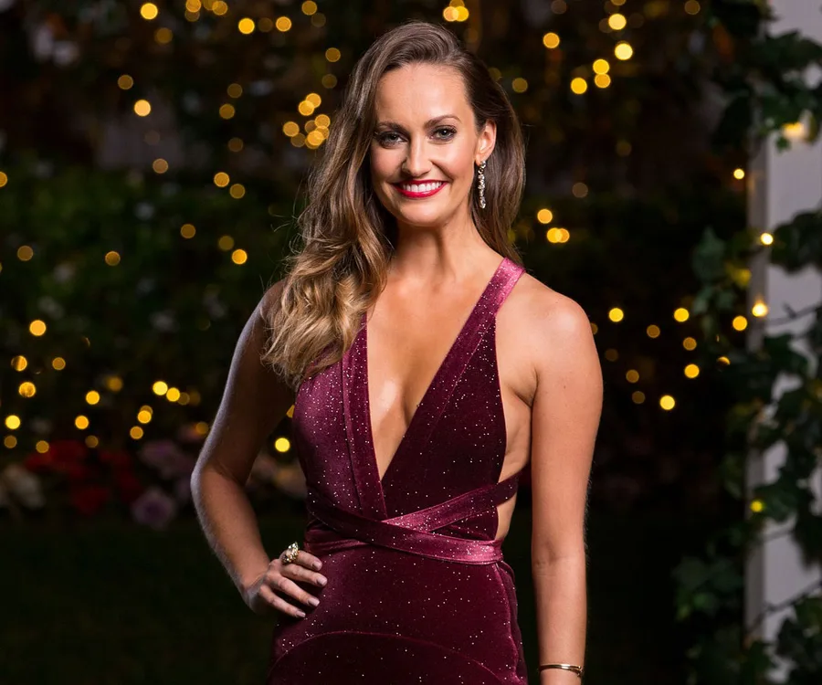 A woman in a sparkly burgundy dress stands smiling with blurred lights in the background.