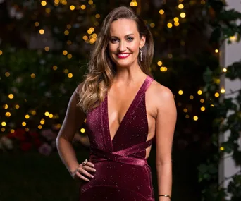 A woman in a sparkly burgundy dress stands smiling with blurred lights in the background.