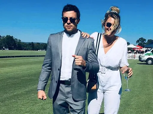 A couple dressed in stylish outfits walk on grass under a clear blue sky, with the woman holding a glass.
