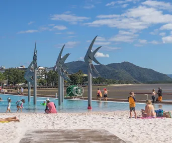 Children and adults enjoying the Lagoon Pool in Cairns with iconic metal fish sculptures and mountains in the background.