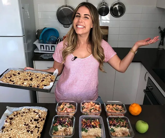 Cooking enthusiast smiling, standing in a kitchen with meal prep containers and holding a tray of granola.