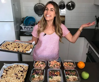 Cooking enthusiast smiling, standing in a kitchen with meal prep containers and holding a tray of granola.