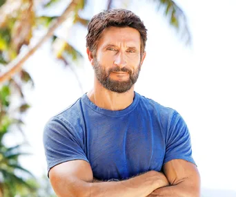 Man with beard in a blue shirt stands with arms crossed, tropical background with palm trees.