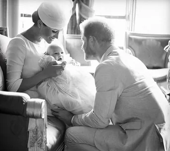 A woman and a man lovingly gaze at a baby swaddled in a blanket, seated in a softly lit room.