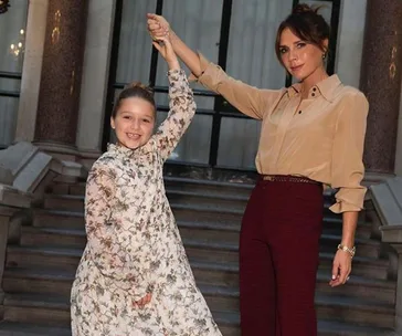Two people pose on steps, holding hands, one in floral dress, one in beige top, smiling at fashion event.