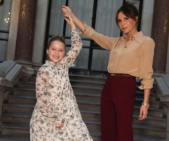 Two people pose on steps, holding hands, one in floral dress, one in beige top, smiling at fashion event.