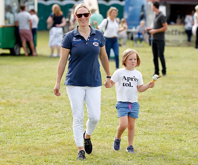 A woman and child holding hands walk on grass. The child has an ice cream cone. Both are smiling outdoors.