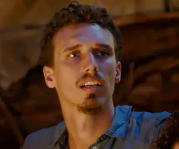 A young man with curly hair and a contemplative expression, set against a dimly lit background.