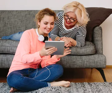 Woman and teen sitting on floor using tablet together, smiling, with headphones visible around teen's neck.