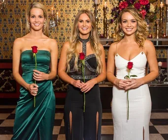 Three women in elegant dresses holding roses, standing in a decorated room.
