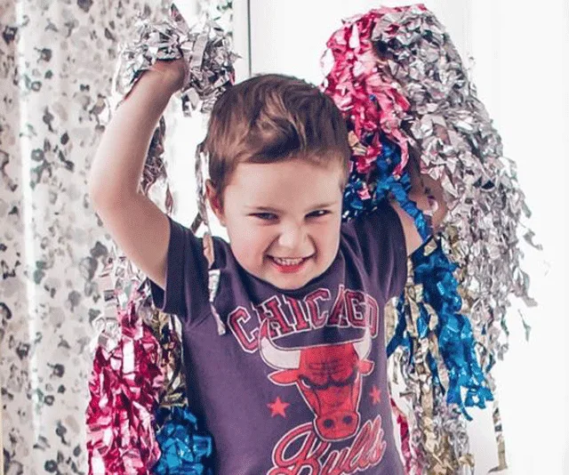 A child with pom-poms smiles, wearing a Chicago Bulls shirt, standing indoors with light filtering through curtains.