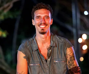 Man with curly hair and arm tattoos, wearing a sleeveless shirt, smiling outdoors at night, with lights blurred in background.