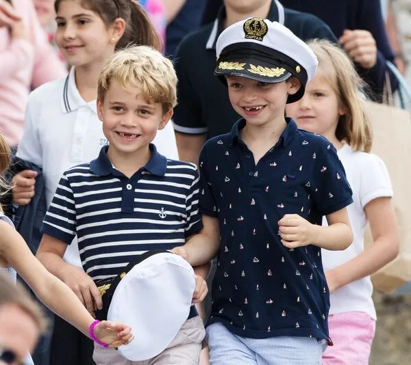 Children smiling and walking together; one wears a navy striped shirt and holds a captain's hat, another wears a captain's hat.