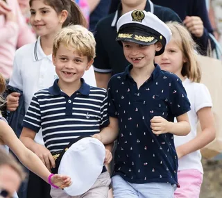 Children smiling and walking together; one wears a navy striped shirt and holds a captain's hat, another wears a captain's hat.