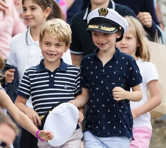 Children smiling and walking together; one wears a navy striped shirt and holds a captain's hat, another wears a captain's hat.
