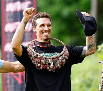 A smiling man with tribal necklace, raising his fist, outdoors.