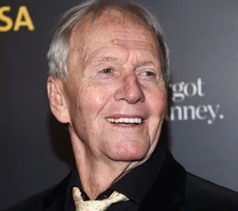 Elderly man smiling at a formal event, wearing a black suit and patterned tie, against a black background.