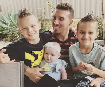 A man smiles with three young children, one in a Pikachu shirt, sitting in a garden setting.