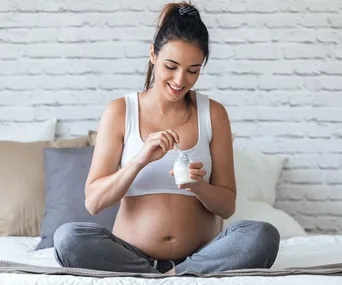 Pregnant woman in casual attire eating yogurt while sitting cross-legged on a bed.